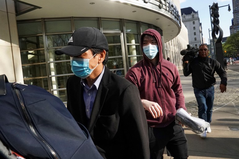 Lu Jianwang lascia il tribunale federale di Brooklyn.  Indossa un berretto da baseball e una maschera facciale ed è seguito da suo figlio che indossa una felpa con il cappuccio alzato e una maschera facciale.