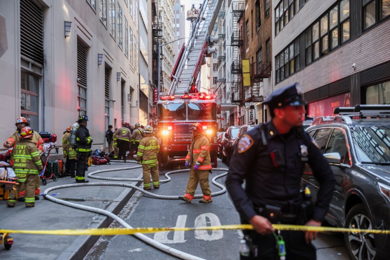 A police officer holds yellow caution tape while firefighters with trucks and ladders work on the narrow street behind him