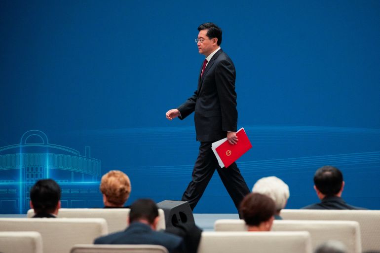 China's foreign minister Qin Gang walks onto a stage in Shanghai. He is carrying a red file in his left hand. The backdrop behind him is blue.