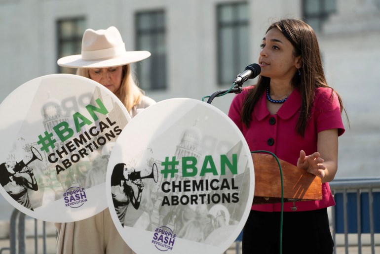 Uma mulher com uma placa redonda que diz "#Proibir abortos químicos" fala em um microfone do lado de fora da Suprema Corte dos EUA.  Há outra mulher ao lado dela, segurando a mesma placa e usando um vestido creme e chapéu.
