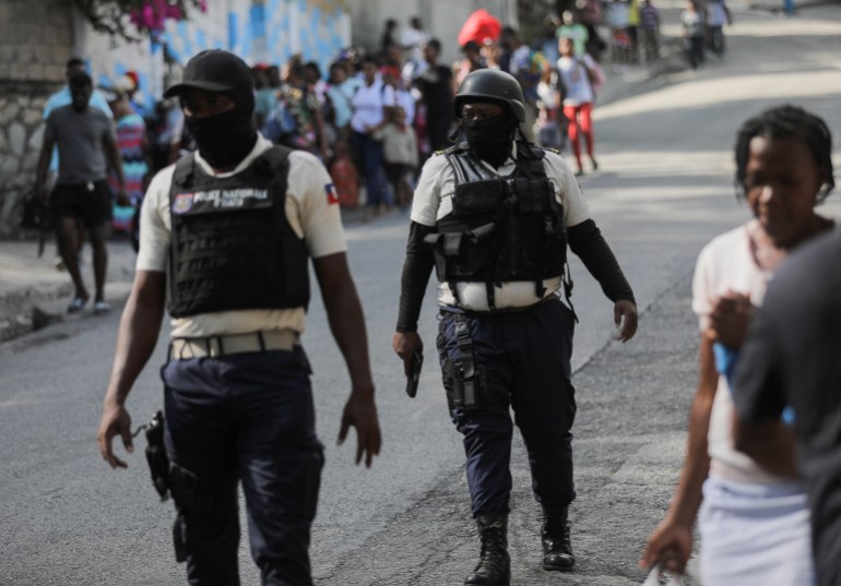 Petugas polisi berjalan di dekat orang-orang yang membawa barang-barang mereka di tengah kekerasan geng di ibu kota Haiti, Port-au-Prince