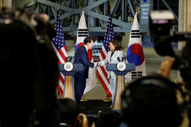 Seen from a crowd, Kamala Harris and Yoon Suk Yeol lean over from their separate podiums to shake hands