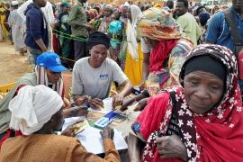 Women getting supplements at a WFP registration desk after fleeing Sudan. They are outside. The woman at the desk has lots of pieces of paper in front of her.