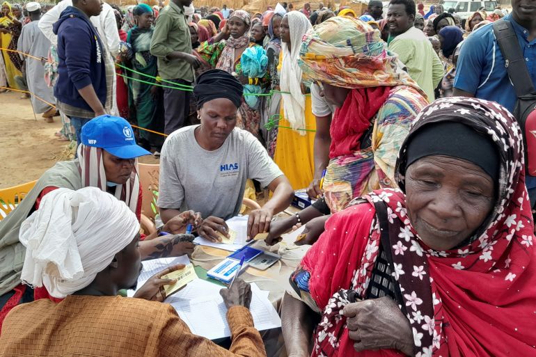 Women getting supplements at a WFP registration desk after fleeing Sudan. They are outside. The woman at the desk has lots of pieces of paper in front of her.
