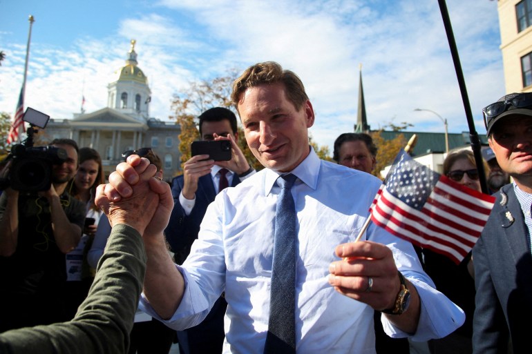 Dean Phillips, wearing a collared shirt and tie, holds a little American flag in one hand and the hand of a supporter in the other.