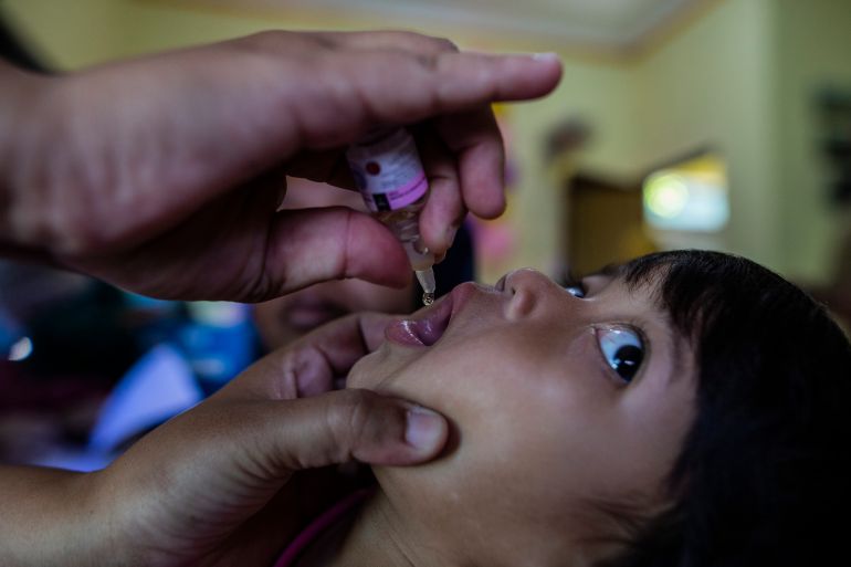 A girl in Indonesia receiving a polio vaccine. She is tipping her head back and looking to the ceiling. The health worker is putting the vaccine on her tongue