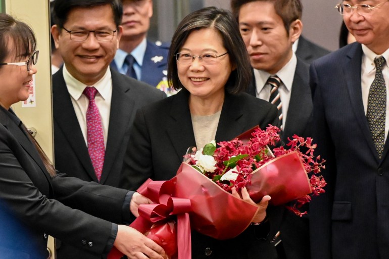 Tsai Ing-wen voltando para casa.  Ela está sorrindo e cercada por funcionários. Ela carrega um buquê de flores vermelhas e brancas que acabaram de ser entregues a ela.