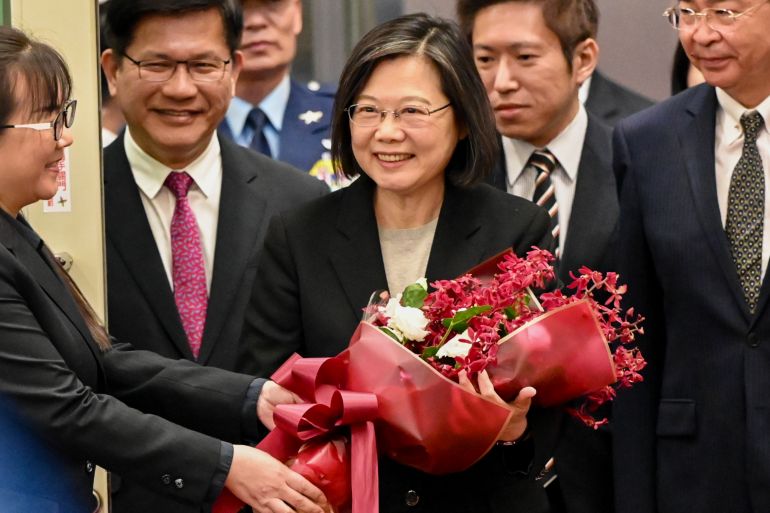 Tsai Ing-wen returning home. She is smiling and surrounded by officials, She is carrying a bouquet of red and white flowers that have just been handed to her.
