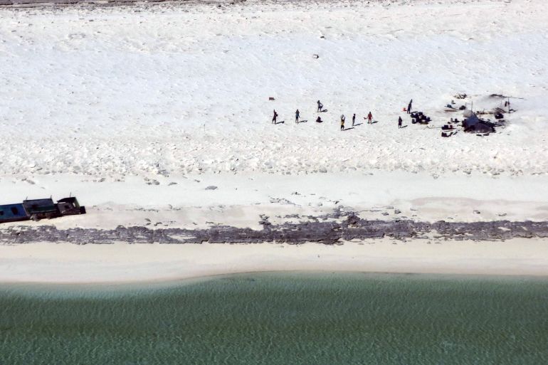 The fishermen standing on the beach of Bedwell Island. Their shipwrecked boat is lying closer to the shore and they have built a makeshift shelter. The picture is taken from the sky.