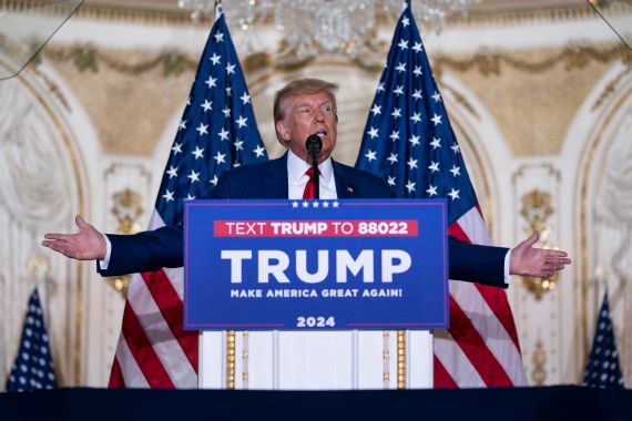 Donald Trump at podium, arms outstretched, speaking in front of two US flags