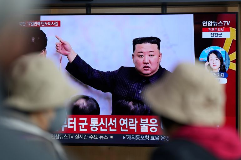People watching a large TV screen of a news programme. On the large screen there is a picture of Kim Jong Un pointing at a map/