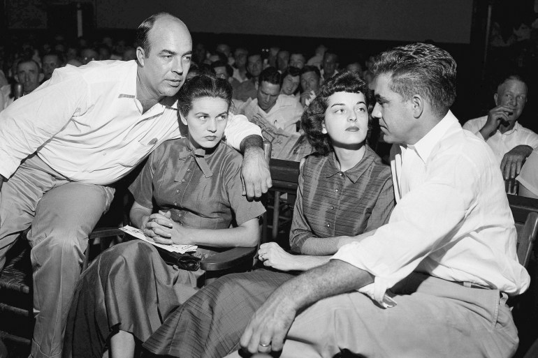 A man with his arm around a seated woman leans over, while another couple is seated to their right.