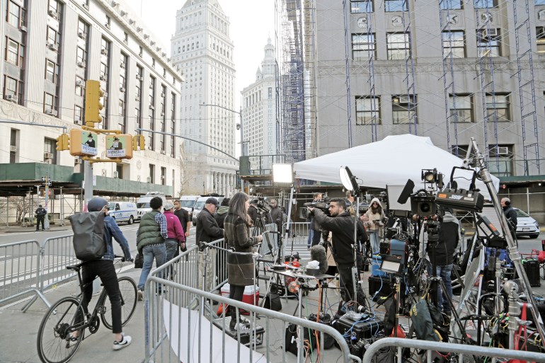 Cameras clustered on a sidewalk in lower Manhattan as reporters do live coverage. They're cordoned off by a low fenced barrier. On the outside, a biker passes through. 
