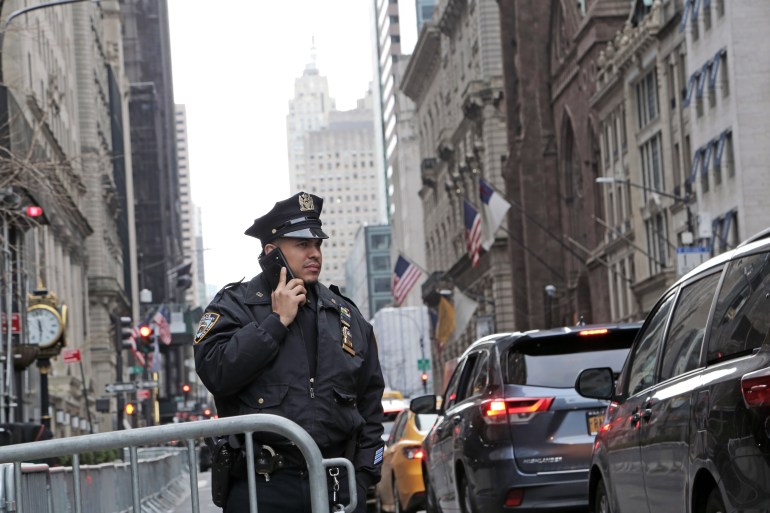 A police officer speaks on the phone in Manhattan traffic. Behind him, US flags hang on slanted poles coming off buildings on either side of the street. 