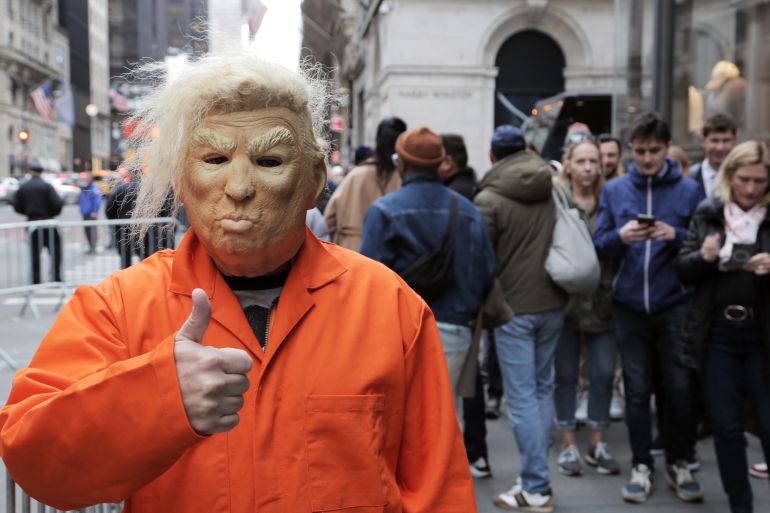 A protester in a Trump mask, with bushy eyebrows, and an orange jumpsuit gives a thumbs-up to the camera. 