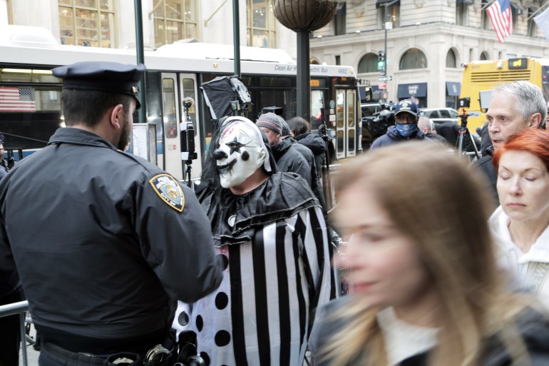 A police officer on a busy sidewalk, speaking to a man dressed as a creepy black-and-white clown. People on the right walk past. There is a blurry close up of a woman in motion.