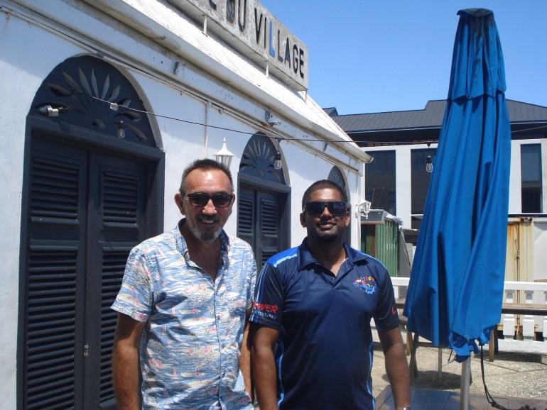Simon Troman and Mark Philips on the waterfront at Port VIla standing outside Troman's cafe. The building's shutters are closed and there is furled blue umbrella to their left.