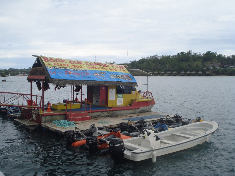 Sebuah ponton terapung dan tiga perahu milik U Power Adventures di tepi pantai Port Vila.  Ada spanduk besar yang menutupi atap yang mengiklankan bisnis tersebut.  Jaket pelampung menggantung.  Langit berawan