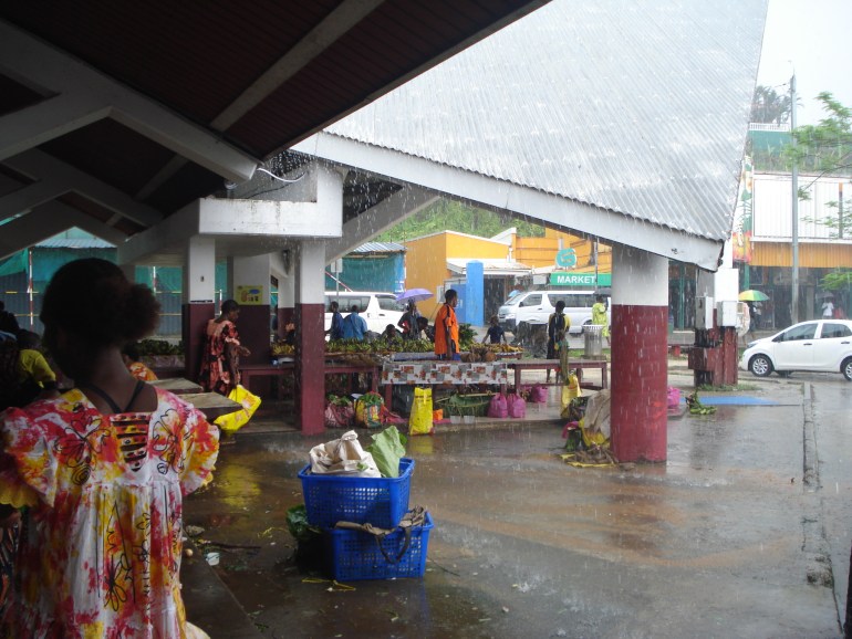 Rain lashes a Vanuatu market. There is a woman to the left with her back to the camera in a colourful, floral dress. There are stalls in the middle undercover with a few people milling about. The ground is very wet and rain is cascading from the roof.