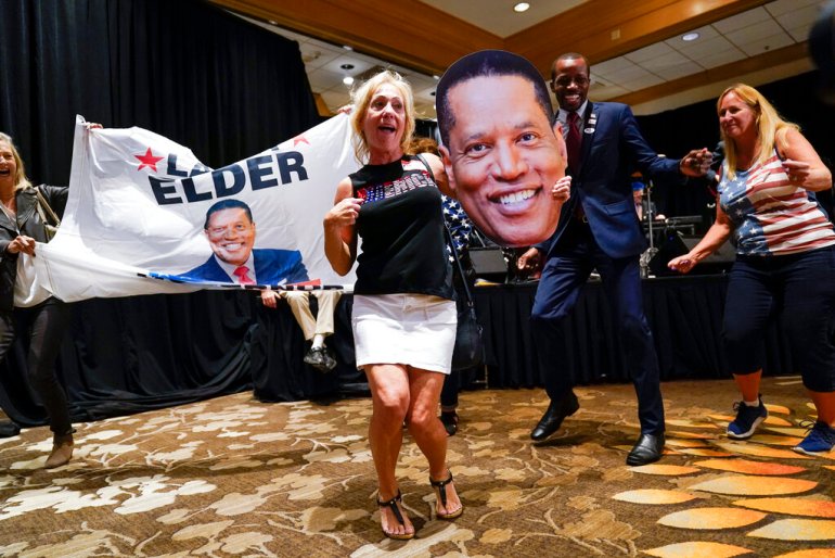 Larry Elder dances with supporters who are holding a campaign banner and a large cutout of his face.