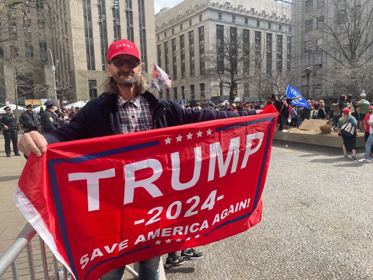 A man in a red cap holds a "Trump 2024" flag outdoors in Manhattan