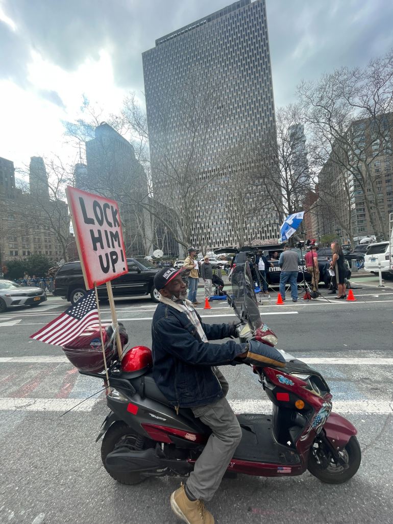 A man sits on a motorbike with an American flag and a protest sign pinned to the back