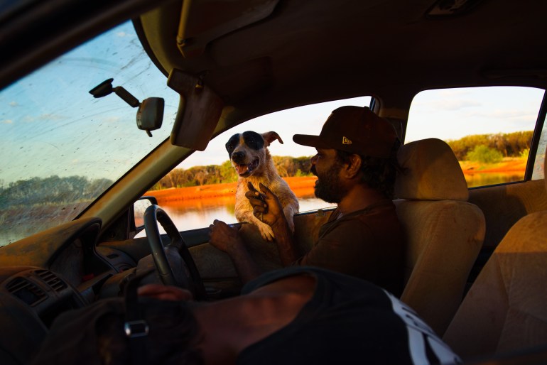 Lloyd Jampijinpa Brown assis du côté conducteur d'une voiture.  Il caresse son chien Blackeye qui se penche vers la voiture de l'extérieur, ses pattes posées sur le rebord de la fenêtre.