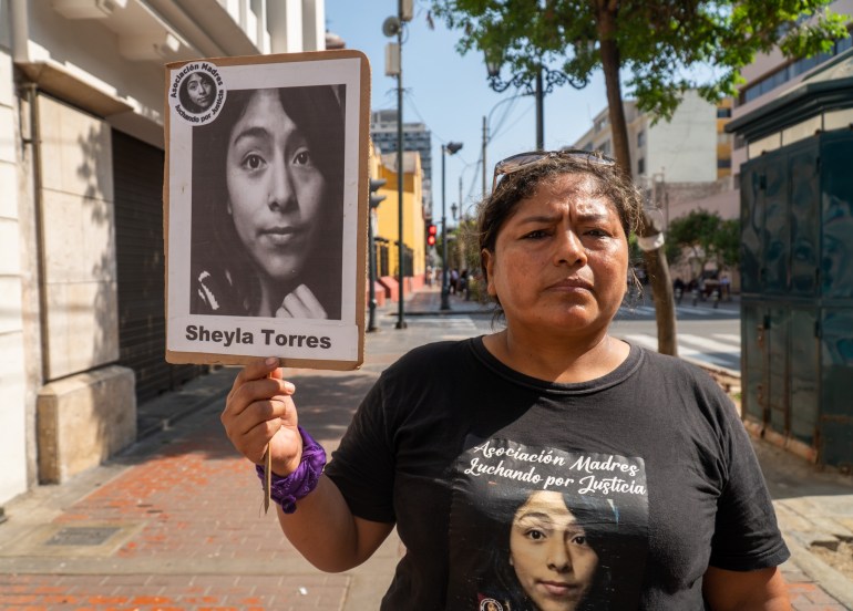 A woman walks down the street holding a sign with the name and portrait of Sheyla Torres on it.  She also wears a t-shirt with Sheila's face on it. 