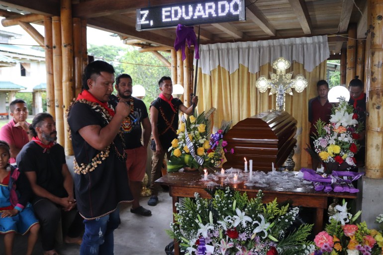 Une personne se tient en prière à côté d'un cercueil décoré de fleurs devant une croix