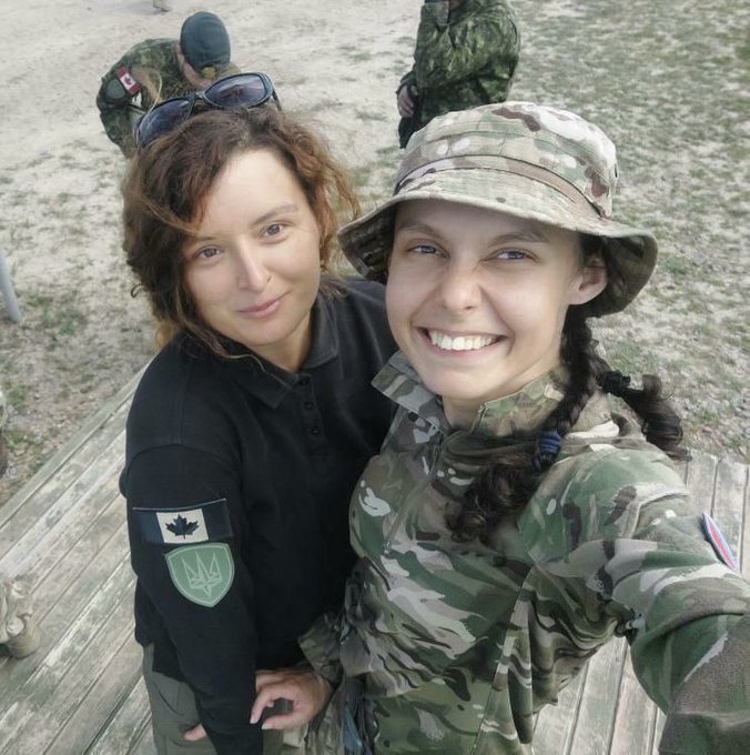 Two women are looking up at the camera. Kudriava is wearing camouflage uniform and hat while Ferlain, with curly red hair, hugs her friend. She is wearing a black jersey.