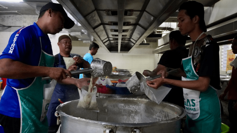 Men fill small plastic packets with piping hot porridge. They are pouring the mixture into the bag with a metal funnel. There is a huge cooking pot between them. Other men are working behind them.
