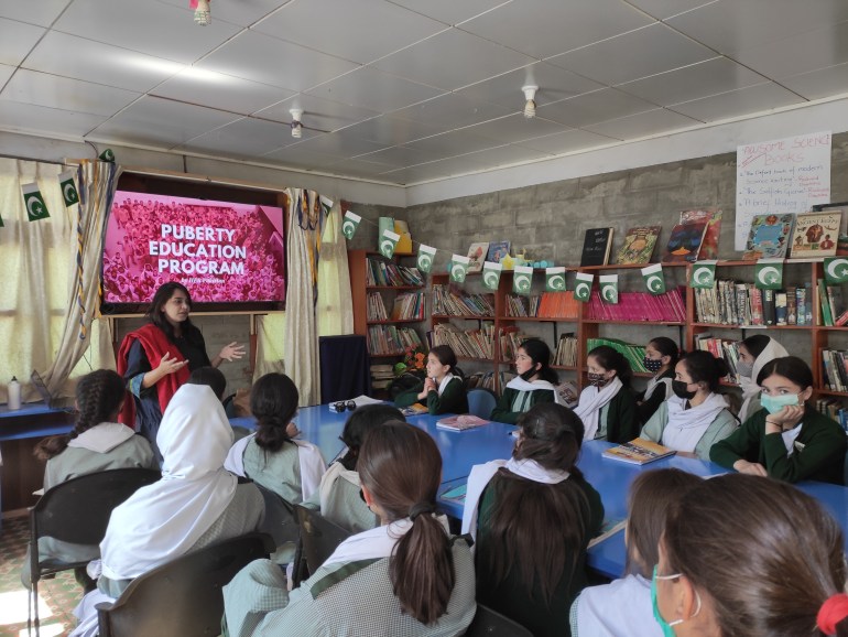 A photo of Sumaira holding a presentation for female students with a television in the background with the words "Puberty education program" on it.