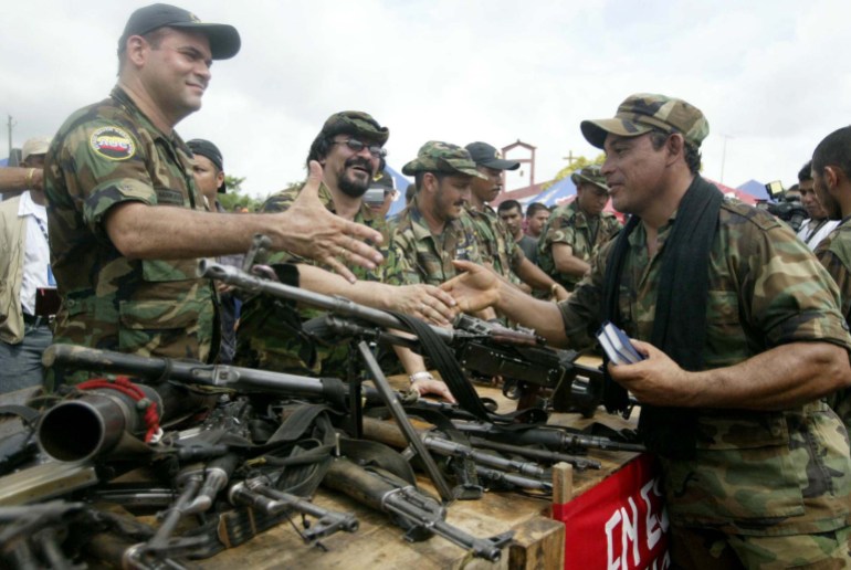 Seorang pria berseragam militer meraih tumpukan senjata api untuk berjabat tangan dengan pria lain yang mengenakan perlengkapan militer, memegang dua buku catatan.  Anggota paramiliter lainnya menyaksikan dari seberang meja dengan senjata api.
