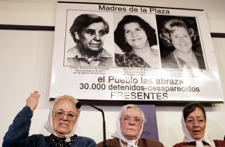 Tiga wanita dengan syal putih di atas kepala mereka duduk di bawah poster dengan potret hitam putih dari tiga wanita yang hilang