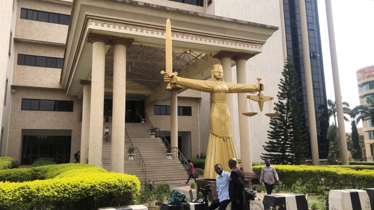 Journalists wait outside the Federal High Court, after the leader of Indigenous People of Biafra (IPOB), Nnamdi Kanu, was brought to court according to government sources, in Abuja, Nigeria. A golden statue of a lady in a blindfold holding scales and a sword sits at the base of the building's steps.