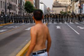 A young man, shirtless, stands in the middle of an empty city road, staring at a line of police officers at the other end of the street.
