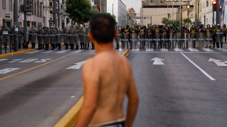 A young man, shirtless, stands in the middle of an empty city road, staring at a line of police officers at the other end of the street.