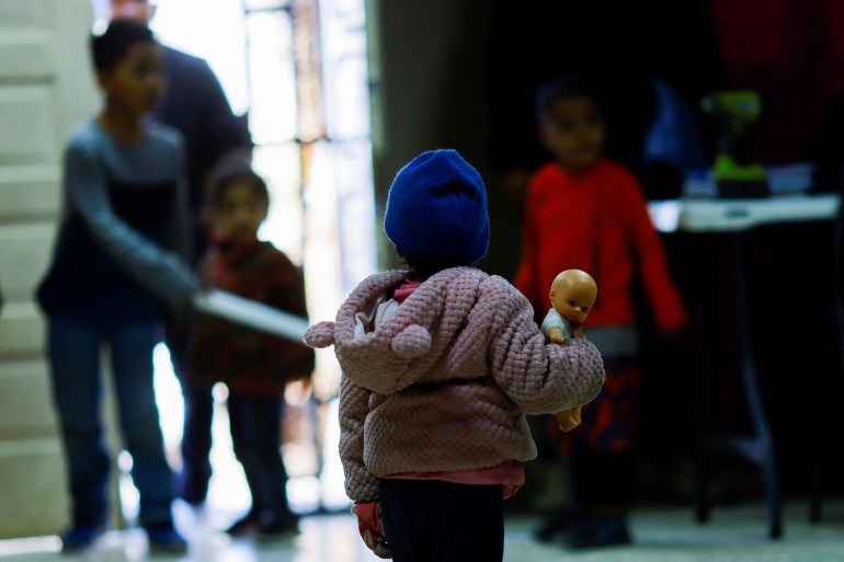 A girl in a beanie hat is seen from behind holding a doll in a shelter room
