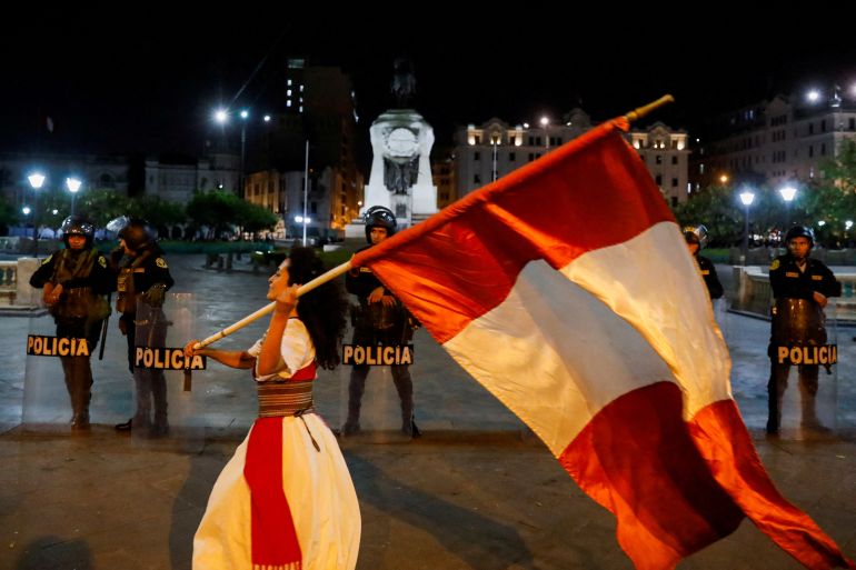 A woman carrying a large Peruvian flag over her shoulder strolls by a line of riot police in Lima