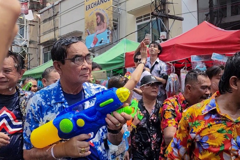 Prayuth Chan-ocha in a blue Hawaiian shirt at Songkran festivities in Bangkok. He has a large blue water gun, Others are spraying him with water and his hair is wet. He is not smiling.