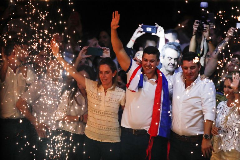 Paraguayan presidential candidate Santiago Pena celebrates his victory. He is standing with his wife, and running mate. They are punching the air. They are surrounded by sparkling light.