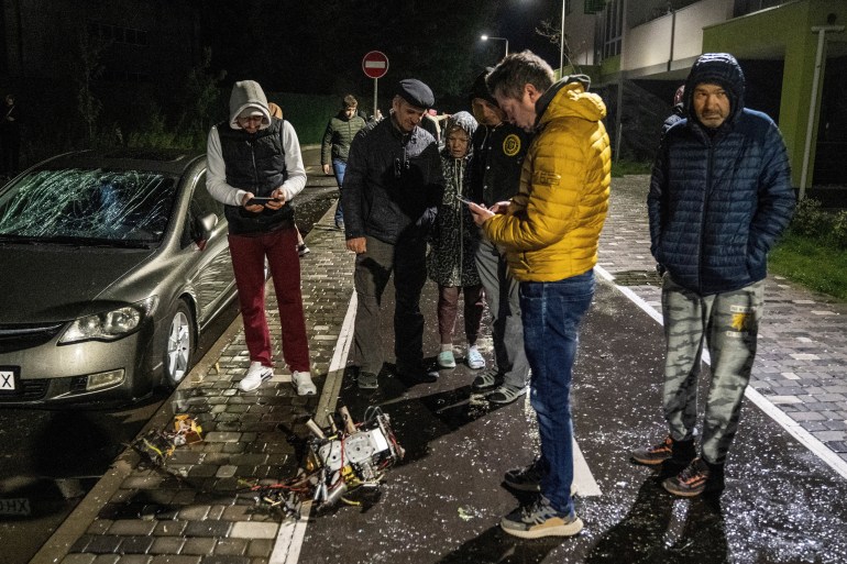 Kyiv residents inspect debris from a drone that fell in their neighbourhood. They are dressed in sweaters and jackets and taking photos of what is left of the drone with their phones. 