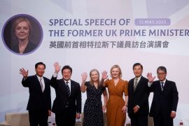 Liz Truss takes a group photo in Taipei. She is waving and smiling. The backdrop behind reads 'Special Speech of the Former UK Prime Minister' with her photo to one side. The words are also written in Chinese.