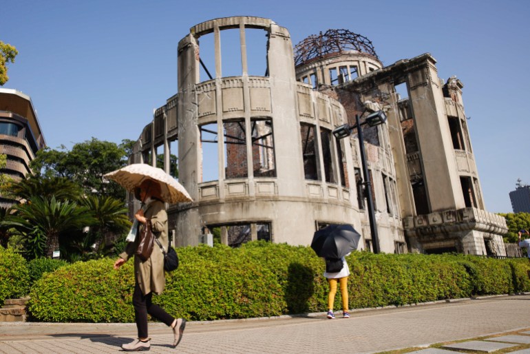 A view of the A-Dome in Hiroshima. The building is in ruins but preserved. Two people are walking in front beneath umbrellas.