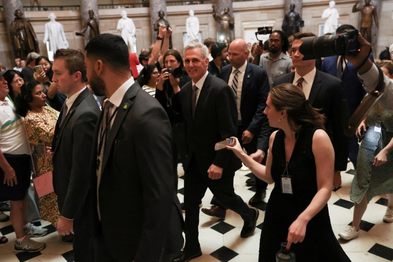 مردی با کت و شلوار تیره از کف کاشی‌شده کنگره ایالات متحده عبور می‌کند، در حالی که افراد دیگری با لباس رسمی احاطه شده‌اند.  مجسمه های سفید رنگ در پس زمینه دیده می شوند.
