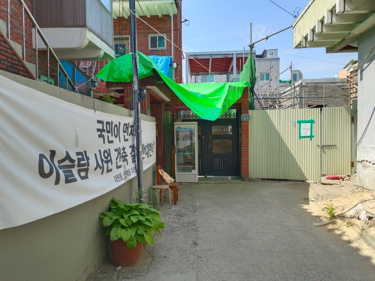 Pathway leading to the mosque construction site. A banner on left reads 'People first! Against the construction of the mosque!' and there is a plastic pig head on a chair in front of it. at the back, next to the site entrance is a refrigerator containing three pig heads. The construction site is on the right. Scaffolding is visible.