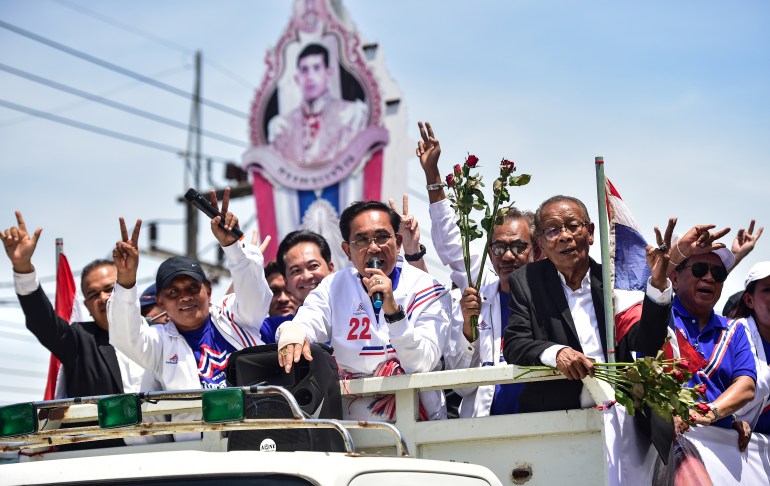 Incumbent prime minister Prayuth Chan-ocha campaigning on the top of a lorry. He is wearing white and surrounded by supporters. There is a picture of the country's king behind him