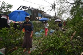 People with umbrellas negotiate trees brought down by Cyclone Mocha in Kyauktaw, Myanmar. There is a building without a roof behind them.