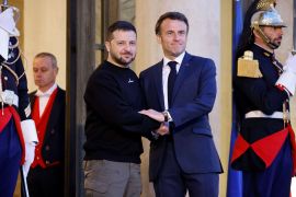 French President Emmanuel Macron greets Ukrainian President Volodymyr Zelenskyy at the Elysee Palace. Macron is in a blue suit and tie, while Zelenksyy is in a black sweath short and khaki trousers. They look resolute.