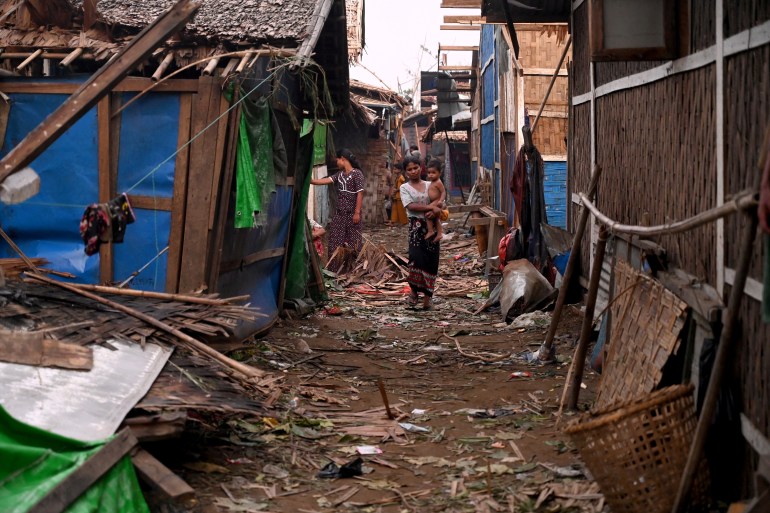 A woman walking through a debris strewn alleyway between bamboo and tarpaulin houses in a Rohingya camp in Sittwe.  She is holding a child.  There is another woman behind her looking inside a building